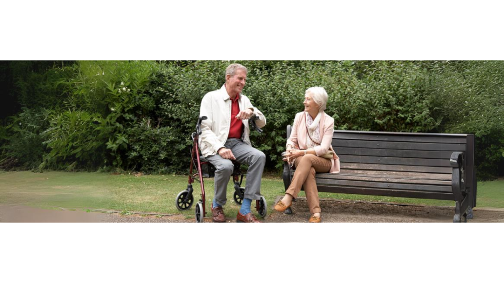 An elderly man seated on a Days Rollator converses with an elderly woman seated on a park bench