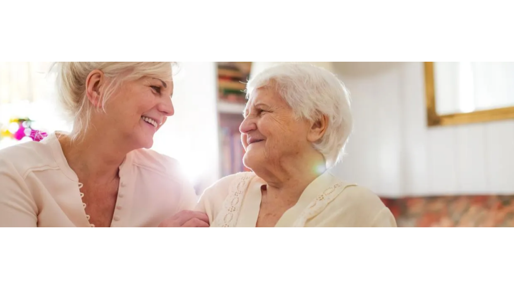 Senior woman & her daughter sitting in living room together, embracing & smiling over tea.